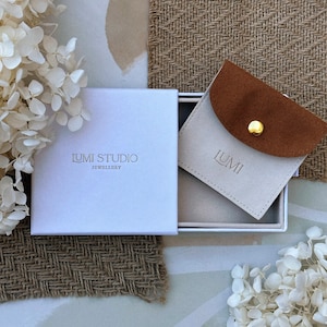 May include: A white box with the text "Lumi Studio Jewellery" on the top. Inside the box is a small, light brown pouch with the text "Lumi" on the front. The pouch is made of suede and has a gold button.