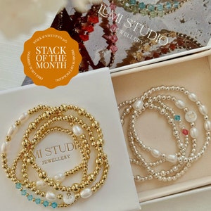 May include: Two sets of bracelets in a white box. One set is gold with white pearls and turquoise beads. The other set is silver with white pearls. Both sets have a letter charm. The box has the text "MI STUDI JEWELLERY" on it.