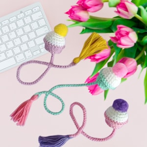 May include: Three crocheted bookmarks with colorful pom-poms and tassels. The bookmarks are pink, green, and purple with white accents. The bookmarks are on a pink background with pink tulips.