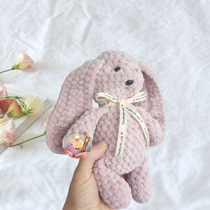 May include: A soft, plush, lavender-colored crocheted bunny rabbit toy with a white ribbon tied around its neck. The ribbon has the text "Handmade with love" printed on it. The bunny is sitting in a hand.