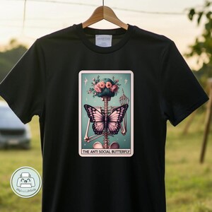 K&ouml;nnte beinhalten: Schwarzes T-Shirt mit einem Grafikdruck eines Skeletts mit einem Schmetterling auf dem R&uuml;cken. Das Skelett tr&auml;gt eine Blumenkrone und zeigt den Mittelfinger. Der Text "The Anti Social Butterfly" befindet sich unter dem Grafikdruck.
