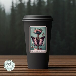 K&ouml;nnte beinhalten: Eine schwarze Kaffeetasse mit einem Aufkleber, der ein Skelett mit einem Schmetterling auf dem R&uuml;cken zeigt. Der Aufkleber tr&auml;gt den Text "The Anti Social Butterfly".