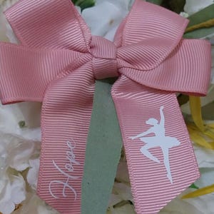May include: A pink ribbon bow featuring a ballerina silhouette and the word "Hope" in white. The bow is made of a ribbed material. The background includes white and yellow flowers.