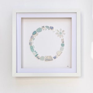 May include: A framed artwork with a white mount and frame. The artwork is a circular arrangement of sea glass and ceramic shards in shades of blue, teal, and white. A flower shape is created with similar pieces. The art is mounted on a white background.
