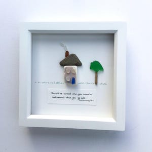 May include: A white framed artwork with a house and tree design crafted from sea glass. The house has a gray roof, a white body, and a blue door. A green tree stands beside it. The text reads, "You will be blessed when you come in and blessed when you go out. Deuteronomy 28:6."