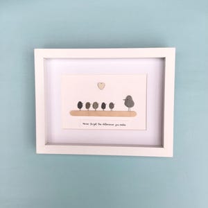 May include: A white framed wall art piece featuring pebble art of five small birds and one larger bird standing on a wooden branch. Above the birds is a small wooden heart. Below, a white label displays the text: Never forget the difference you make.