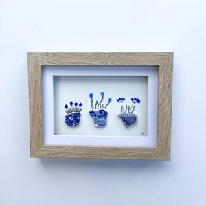 May include: A framed artwork featuring three blue and white floral arrangements. The artwork is composed of pieces of broken pottery arranged to resemble potted plants with blue flowers. The frame is made of light-colored wood.