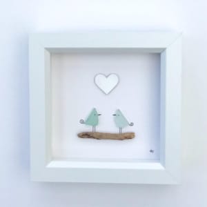 Peut inclure: Une œuvre d'art encadrée blanche représentant deux oiseaux stylisés bleu-vert sur un morceau de bois flotté, avec un cœur blanc au-dessus. Le cadre carré met en valeur le design minimaliste et moderne.