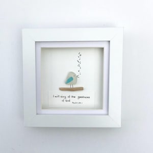 May include: A framed artwork depicting a small, pale bird with blue detailing, perched on a wooden branch. The image includes musical notes and the text "I will sing of the goodness of God" with a Bible verse reference. The frame is white.