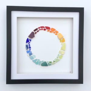 May include: Framed artwork with a circular design made from colourful sea glass pieces. The sea glass is arranged in a spectrum of colours, from red to blue, within a white mount and black frame. A unique piece of wall decor.