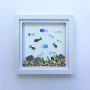 May include: A framed artwork featuring a whimsical underwater scene. Colorful glass fish of various sizes and designs swim above a bed of pebbles, seaweed, and small rocks. The artwork is encased in a white square frame.