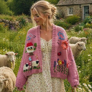 Op de afbeelding: Een roze vest met borduursels van een huis, schapen, bloemen en een lieveheersbeestje. Het vest wordt gedragen over een jurk met bloemenprint. Schapen grazen in een veld, met een huisje op de achtergrond.