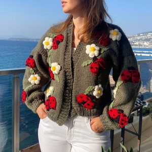 May include: Olive green knit cardigan adorned with red ladybugs and white daisy appliqu&eacute;s. The open-front sweater features long sleeves and a relaxed fit. The ladybugs have black spots and the daisies have yellow centers. The cardigan is paired with white pants.