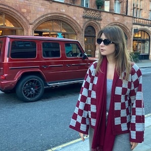 Könnte beinhalten: Eine Person trägt ein graues Oberteil, eine graue Hose, eine schwarze Sonnenbrille, einen bordeauxfarbenen Schal und eine grau-bordeauxfarbene karierte Jacke. Ein roter SUV parkt im Hintergrund auf der Straße. Die Person steht auf einem Bürgersteig vor einem Gebäude.