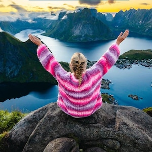 May include: A person wearing a pink and white striped sweater with their arms raised, sitting on a rocky cliff overlooking a body of water. The water is surrounded by mountains and a small town.