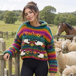 Op de afbeelding: Een kleurrijke trui met capuchon en een regenboogstrepenpatroon. De trui heeft witte schapen en gele bloemenapplicaties. De persoon draagt ​​zwarte leggings en groene regenlaarzen. De achtergrond toont een houten hek, schapen en een paard.