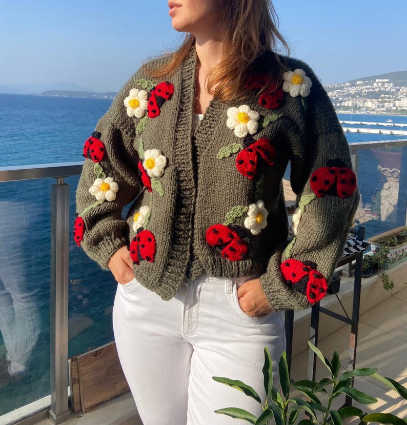 May include: Olive green knit cardigan adorned with red ladybugs and white daisy appliqu&eacute;s. The open-front sweater features long sleeves and a relaxed fit. The ladybugs have black spots and the daisies have yellow centers. The cardigan is paired with white pants.