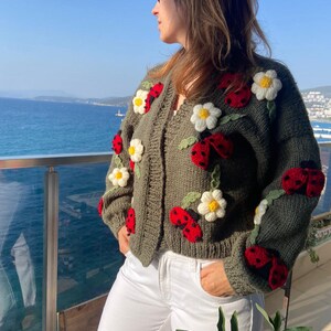 May include: Olive green cardigan sweater adorned with red ladybugs and white daisy flower appliqu&eacute;s. The open-front sweater is a chunky knit with a V-neck design. The ladybugs have black spots and the daisies have yellow centers.