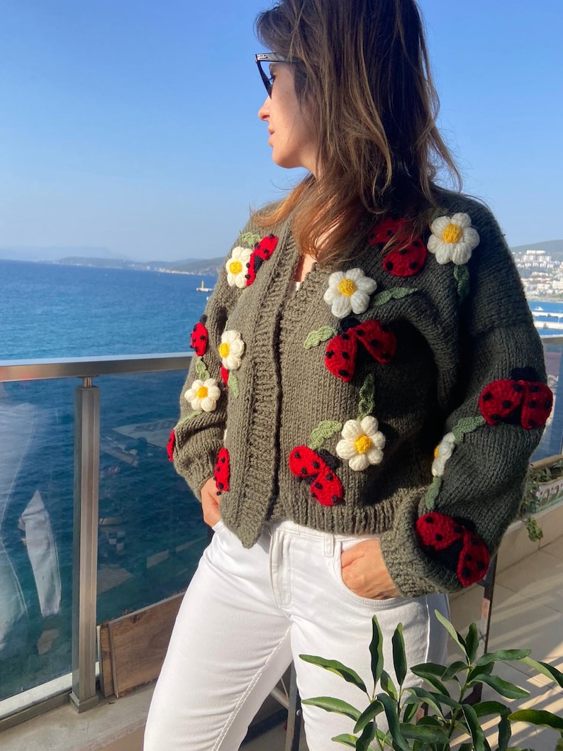 May include: Olive green cardigan sweater adorned with red ladybugs and white daisy flower appliqu&eacute;s. The open-front sweater is a chunky knit with a V-neck design. The ladybugs have black spots and the daisies have yellow centers.