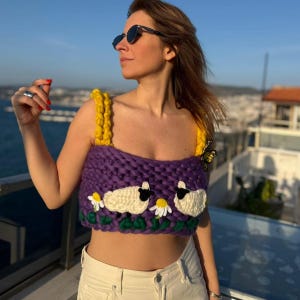 Puede incluir: Un top corto de crochet morado con tirantes amarillos, con detalles de ovejas blancas y margaritas. El top se usa con jeans blancos y gafas de sol. Una mariposa está sujeta al top.