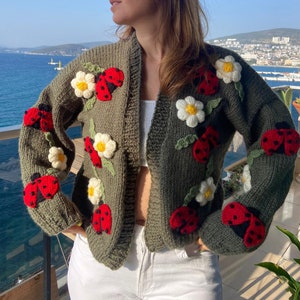 May include: Olive green knit cardigan adorned with red ladybugs and white daisy appliqu&eacute;s. The open-front sweater features long sleeves and is a cozy, handmade garment. The ladybugs have black spots and the daisies have yellow centers.