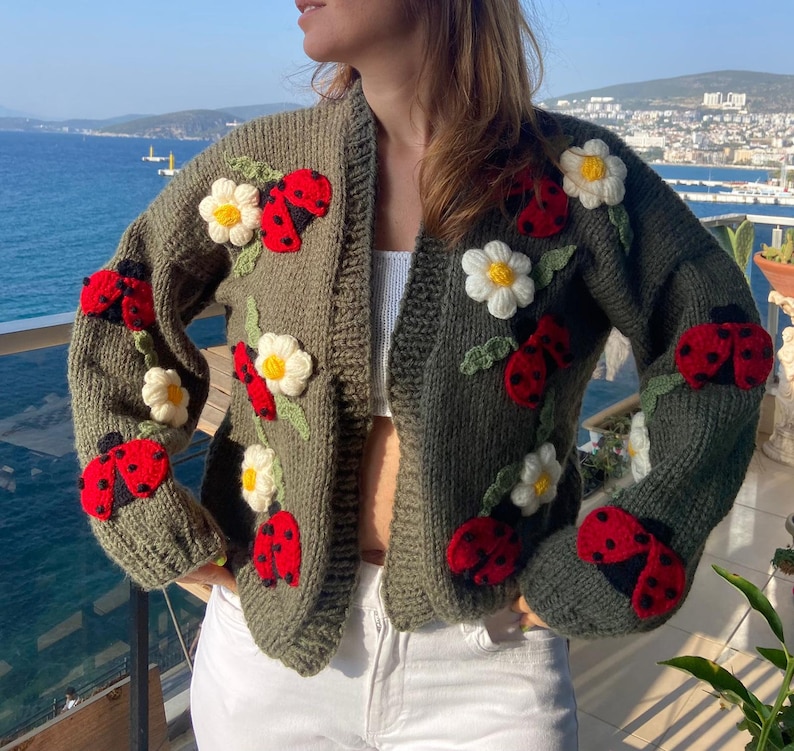 May include: Olive green knit cardigan adorned with red ladybugs and white daisy appliqu&eacute;s. The open-front sweater features long sleeves and is a cozy, handmade garment. The ladybugs have black spots and the daisies have yellow centers.