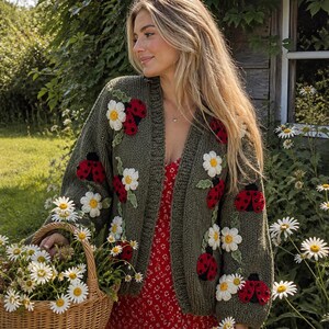 May include: An olive green cardigan adorned with red ladybugs and white daisy appliqu&eacute;s. The open-front sweater is worn over a red floral dress. A woven basket filled with daisies is held in the foreground, suggesting a garden theme.