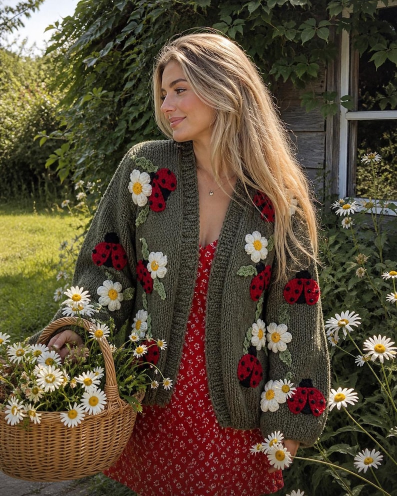 May include: An olive green cardigan adorned with red ladybugs and white daisy appliqu&eacute;s. The open-front sweater is worn over a red floral dress. A woven basket filled with daisies is held in the foreground, suggesting a garden theme.