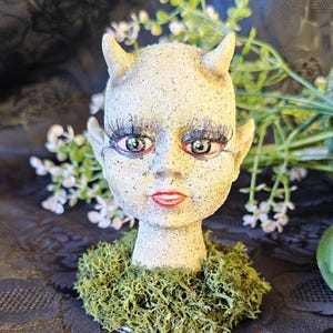 May include: A grey, sculpted head with horns, green eyes, and red lips. The head is decorated with moss and is a decorative piece.