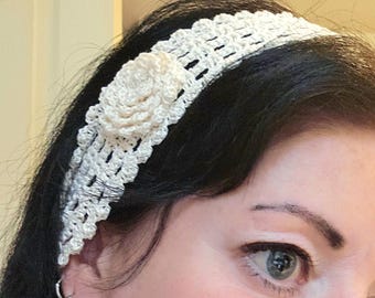 May include: A cream-colored crochet headband with a floral accent. The headband is made of a delicate, open-weave crochet pattern. A cream-colored, three-dimensional flower is attached to the side of the headband. The headband is worn on a person's head.