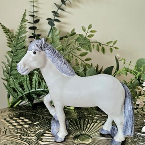 Puede incluir: Una figura de caballo blanco con crin y cola de color gris texturizado. El caballo se muestra en posición de pie con un casco delantero levantado, apoyado sobre una pequeña roca gris. La figura presenta un cuerpo blanco liso y detalles grises.