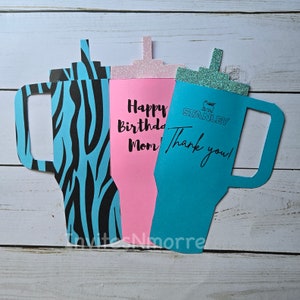 Pode incluir: Três cartões em forma de copo de papel com uma alça. O cartão à esquerda é azul e preto com estampa de zebra. O cartão do meio é rosa com o texto "Happy Birthday Mom". O cartão à direita é azul com o texto "Stanley" e "Thank you!"