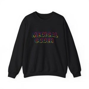 Puede incluir: Sudadera negra con la frase "MEDICAL CODER" en letras de burbujas de colores del arcoíris. La sudadera es de cuello redondo y parece estar hecha de un material suave. El texto está centrado en el pecho.