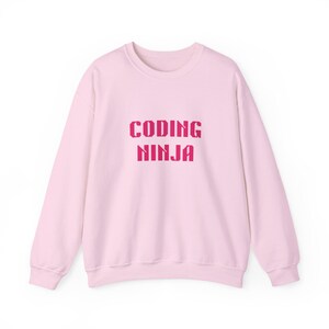 Puede incluir: Una sudadera de manga larga de color rosa claro con el texto "CODING NINJA" impreso en una fuente pixelada de color rosa oscuro en el centro del pecho. La prenda tiene cuello redondo, puños acanalados y un dobladillo acanalado a juego.
