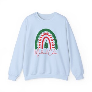 Puede incluir: Sudadera azul claro con un diseño de arcoíris navideño. El arcoíris presenta colores verdes, rojos y blancos, con un árbol de Navidad en el centro. Las palabras "Medical Coder" están escritas en rojo debajo.