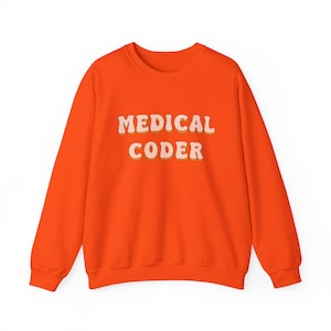 May include: Bright orange sweatshirt with the text "MEDICAL CODER" in a retro font. The text is in white with a light orange outline. The sweatshirt has long sleeves and a crew neck.