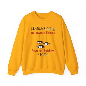 Può includere: Felpa giallo senape con la scritta "Medical Coding Halloween Edition" e "Fear of Spiders F40.210". Il design include tre ragni dei cartoni animati appesi a dei fili.