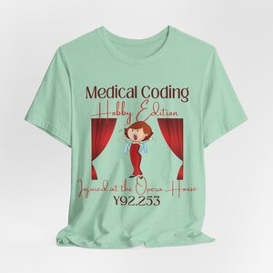 Peut inclure: T-shirt vert clair avec l'inscription "Medical Coding Hobby Edition" et "Injured at the Opera House". Le motif comprend un dessin animé d'une femme en robe rouge sur une scène avec des rideaux rouges.