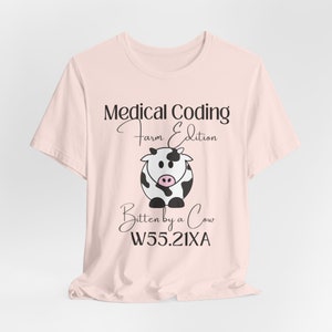 Peut inclure: T-shirt rose clair avec l'inscription "Medical Coding Farm Edition" et une illustration de vache de dessin animé. En dessous, le texte "Bitten by a Cow W55.21XA". La vache est noire et blanche avec un museau rose.