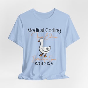 Op de afbeelding: Lichtblauw T-shirt met de tekst "Medical Coding Farm Edition" en een cartoon gans illustratie. De tekst "Bitten by a Goose" en de code "W61.51XA" staan ook op het shirt.
