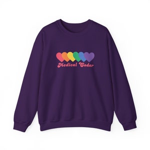 Puede incluir: Una sudadera morada oscura con una fila de seis corazones coloridos en colores del arcoíris. El texto "Medical Coder" está impreso debajo de los corazones en una fuente rosa retro. La sudadera es de color morado oscuro liso.