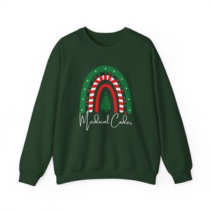 Puede incluir: Sudadera verde oscuro con un diseño de arcoíris navideño. El arcoíris tiene un árbol de Navidad en el centro, con estrellas blancas y rayas rojas y blancas. Las palabras "Medical Coder" están escritas en blanco debajo.