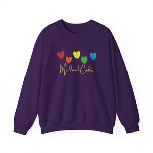 Peut inclure: Un sweat-shirt violet avec une rangée de six cœurs aux couleurs de l'arc-en-ciel. En dessous des cœurs, les mots "Medical Coder" sont écrits en cursive jaune doré. Le sweat-shirt a un col rond et des manches longues.