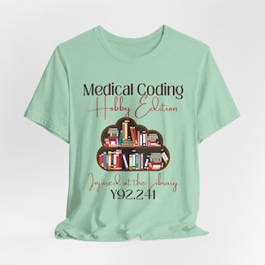 以下が含まれることがあります： 薄い緑色のTシャツに「Medical Coding Hobby Edition」という文字と、雲の中に本棚と本が描かれたグラフィックがあります。「Injured at the Library Y92.241」という文字がグラフィックの下にあります。