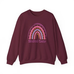 May include: Burgundy sweatshirt with a rainbow design and the text "Medical Coder". The rainbow features pink, purple, and gold colours. The sweatshirt is a long-sleeved crew neck.