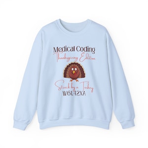 Op de afbeelding: Lichtblauw sweatshirt met de tekst "Medical Coding Thanksgiving Edition" en een cartoon kalkoen illustratie. Onder de kalkoen staat de tekst "Struck by a Turkey" en een code. Een feestelijk kledingstuk voor Thanksgiving.