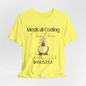Puede incluir: Camiseta amarilla con el texto "Medical Coding Farm Edition" y una ilustración de un pato. Debajo del pato está el texto "Struck by a Duck W61.62XA". La camiseta es de material suave.