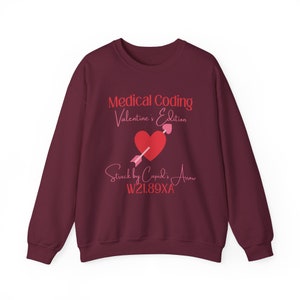 May include: Burgundy sweatshirt with the text "Medical Coding Valentine's Edition" above a red heart pierced by a pink arrow. Below the heart is the text "Struck by Cupid's Arrow" and the code W21.89XA.