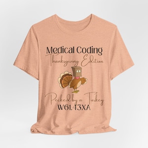 Op de afbeelding: Een licht perzikkleurig T-shirt met de tekst "Medical Coding Thanksgiving Edition" en een cartoon-kalkoen met een papieren zak over zijn kop. De tekst "Pecked by a Turkey W61.43XA" staat onder de kalkoen.