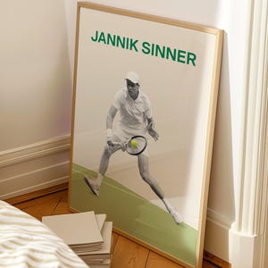 May include: A framed vertical poster featuring a black and white photograph of tennis player Jannik Sinner in a dynamic pose on a green court. The text JANNIK SINNER is printed in bold green capital letters at the top of the white background.